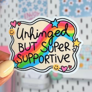 May include: A die-cut sticker with a wavy border featuring a rainbow background and the black text "Unhinged BUT SUPER SUPPORTIVE". The design includes small colorful icons of a yellow smiley face, pink hearts, and blue and yellow stars.