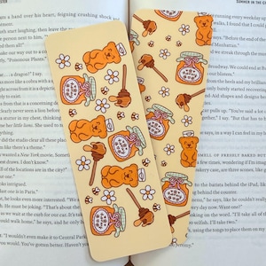 May include: Two rectangular bookmarks with a repeating pattern of honey jars, honey dippers, teddy bears, and flowers on a light yellow background. The bookmarks are lying on top of an open book.