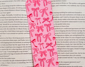 May include: Pink bookmark with a repeating pattern of pink bows. The bookmark is rectangular and is placed on top of a book with printed text.