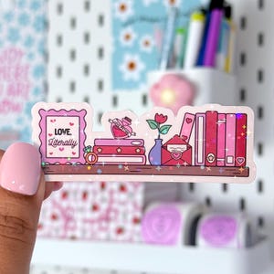 Peut inclure: Un autocollant holographique repr&eacute;sentant une mini-biblioth&egrave;que avec des &eacute;l&eacute;ments romantiques. L'autocollant comprend une image encadr&eacute;e avec le texte "LOVE, Literally", une pile de livres, une bouteille de potion d'amour, une rose dans un vase et une enveloppe.