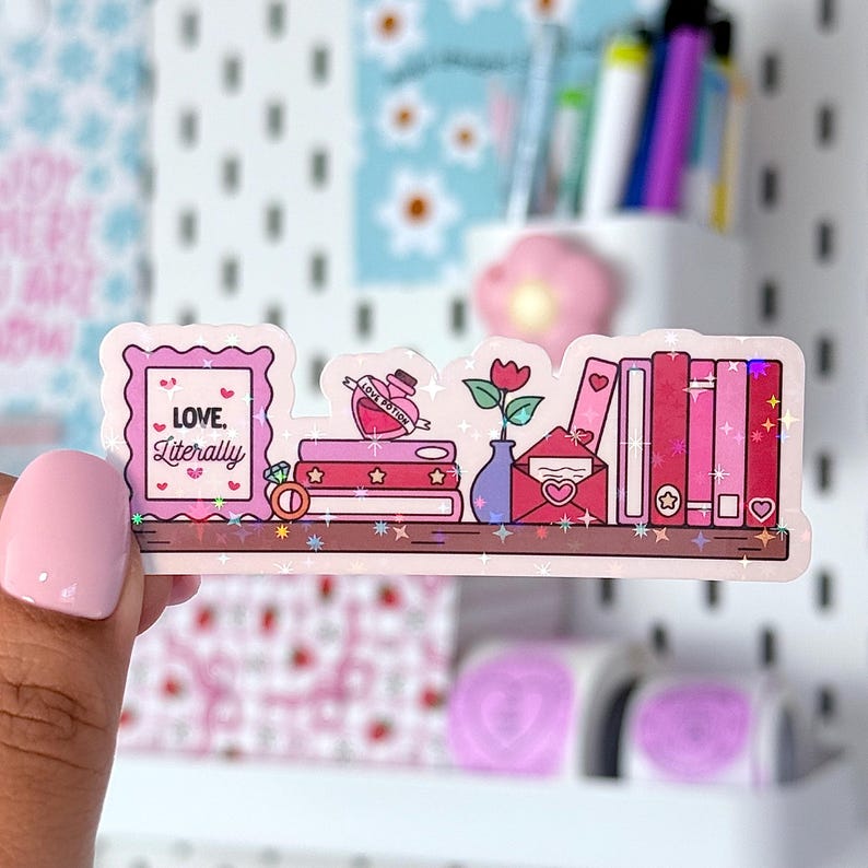Peut inclure: Un autocollant holographique repr&eacute;sentant une mini-biblioth&egrave;que avec des &eacute;l&eacute;ments romantiques. L'autocollant comprend une image encadr&eacute;e avec le texte "LOVE, Literally", une pile de livres, une bouteille de potion d'amour, une rose dans un vase et une enveloppe.