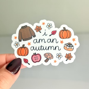 Puede incluir: Una pegatina troquelada con el texto "i am an autumn" rodeada de ilustraciones de un su&eacute;ter de punto marr&oacute;n, calabazas naranjas, una manzana de cuadros rojos, una seta, hojas de oto&ntilde;o, peque&ntilde;as flores blancas y estrellas naranjas.