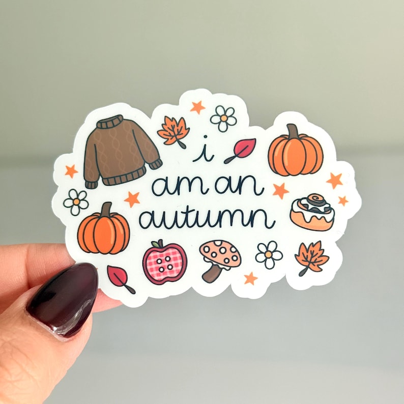 Puede incluir: Una pegatina troquelada con el texto "i am an autumn" rodeada de ilustraciones de un su&eacute;ter de punto marr&oacute;n, calabazas naranjas, una manzana de cuadros rojos, una seta, hojas de oto&ntilde;o, peque&ntilde;as flores blancas y estrellas naranjas.