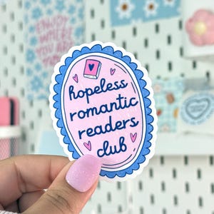 Peut inclure: Un autocollant ovale rose avec une bordure festonn&eacute;e bleue. Le texte "hopeless romantic readers club" est au centre, avec de petites illustrations de c&oelig;urs. Une petite illustration de livre est en haut.