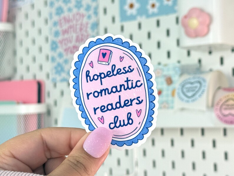 Peut inclure: Un autocollant ovale rose avec une bordure festonn&eacute;e bleue. Le texte "hopeless romantic readers club" est au centre, avec de petites illustrations de c&oelig;urs. Une petite illustration de livre est en haut.