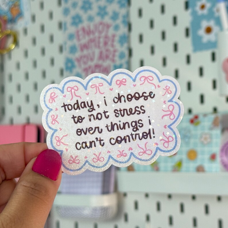May include: A white sticker with a scalloped edge and a light blue border. The sticker has the text "today, I choose to not stress over things I can't control!" in a handwritten style. Pink bows and glitter accents decorate the sticker.