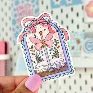 May include: A whimsical sticker featuring a pink butterfly sword piercing an open book. The design includes a pink bow, floral accents, and a light blue scalloped border. The sticker is held by a hand.