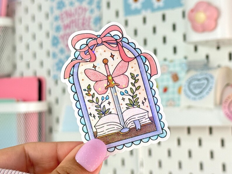 May include: A whimsical sticker featuring a pink butterfly sword piercing an open book. The design includes a pink bow, floral accents, and a light blue scalloped border. The sticker is held by a hand.