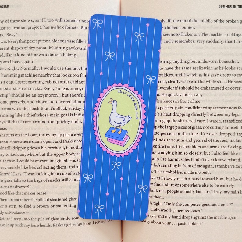 May include: Blue bookmark with a pattern of vertical stripes, bows, and stars. The center features a circular emblem with a goose standing on books, with the text "Silly Goose Book Club".