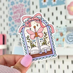 May include: A sticker featuring a whimsical design with a pink butterfly sword piercing an open book. The sticker has a light blue scalloped border and a pink ribbon at the top. Green leaves and stars surround the central image.