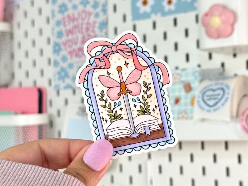 May include: A sticker featuring a whimsical design with a pink butterfly sword piercing an open book. The sticker has a light blue scalloped border and a pink ribbon at the top. Green leaves and stars surround the central image.