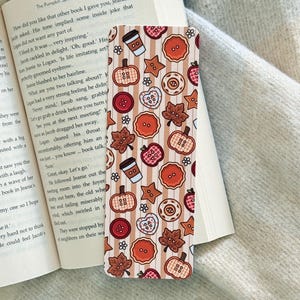 May include: A rectangular bookmark featuring a vertical striped pattern in shades of beige and white. The design includes repeating illustrations of pumpkins, autumn leaves, coffee cups, stars, and circular buttons in red, orange, and brown.