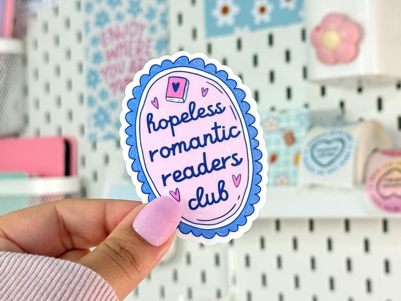 May include: A pink and blue sticker with the words 'hopeless romantic readers club'. The text is in a white, cursive font. The sticker has a blue scalloped border and small heart illustrations. A small pink book illustration is at the top.