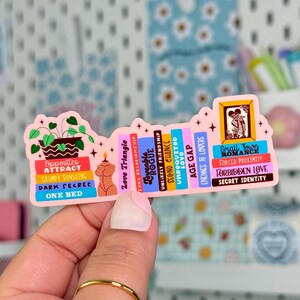 May include: A pink sticker in the shape of a bookshelf, featuring various book titles like "Opposites Attract" and "Forbidden Love." The sticker includes illustrations of plants, a candle, and a framed image of a couple.