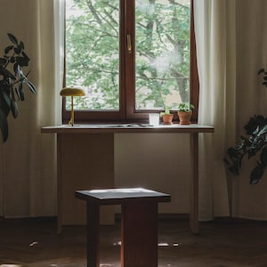 May include: A wooden table with a light wood top and a dark wood base. A small wooden stool sits in front of the table. A yellow lamp sits on the table, and there are two potted plants and a glass of water on the table.