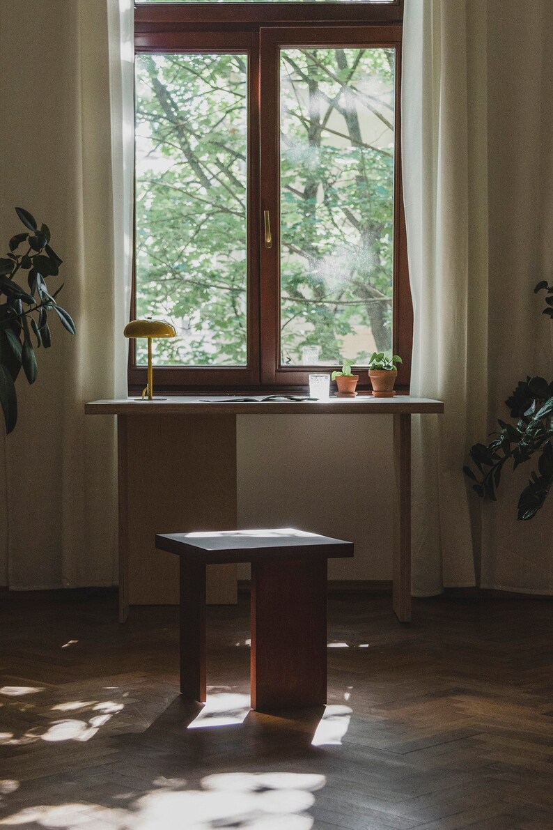 May include: A wooden table with a light wood top and a dark wood base. A small wooden stool sits in front of the table. A yellow lamp sits on the table, and there are two potted plants and a glass of water on the table.