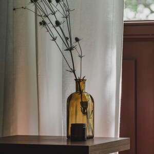 May include: A brown glass bottle with a dried flower arrangement sits on a dark wood table in front of a white sheer curtain.