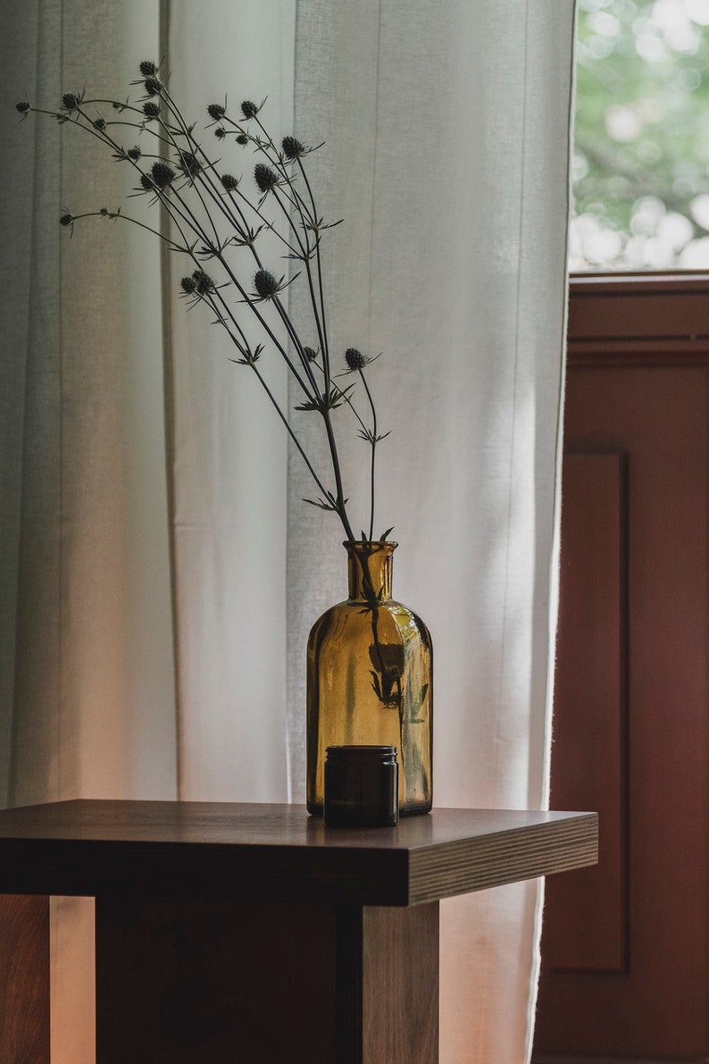 May include: A brown glass bottle with a dried flower arrangement sits on a dark wood table in front of a white sheer curtain.
