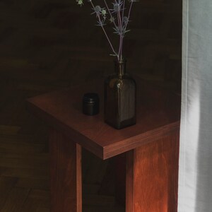May include: A dark brown wooden side table with a single leg on each side. A dark glass bottle with a bouquet of dried thistle flowers sits on the table.