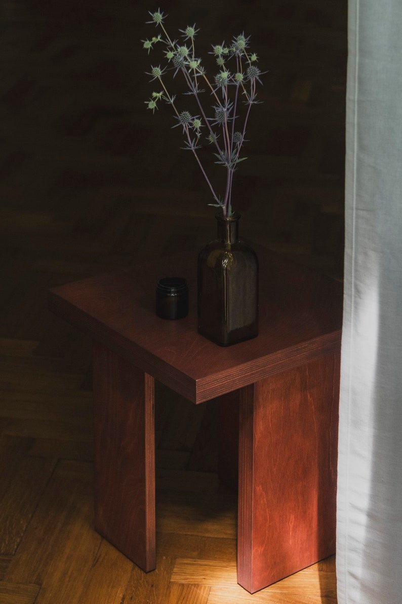 May include: A dark brown wooden side table with a single leg on each side. A dark glass bottle with a bouquet of dried thistle flowers sits on the table.