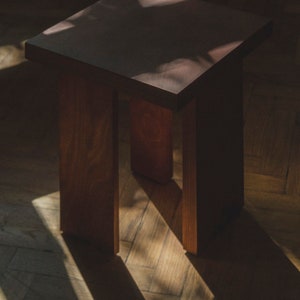 May include: A dark brown wooden stool with a square top and a simple design. The stool is sitting on a wooden floor with light streaming through a window.