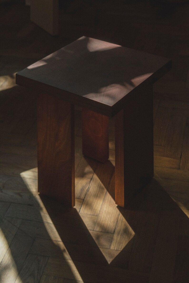 May include: A dark brown wooden stool with a square top and a simple design. The stool is sitting on a wooden floor with light streaming through a window.