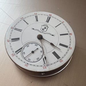 以下が含まれることがあります： 白い背景に黒いローマ数字と小さな秒針が付いた、アンティークの懐中時計の文字盤。 文字盤は金属製のケースに入っており、「THE QUEER WATCH CO.」の文字と航海のエンブレムが表示されています。