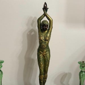 May include: A green and gold statue of a woman with her arms raised above her head. The statue is standing on a black base.