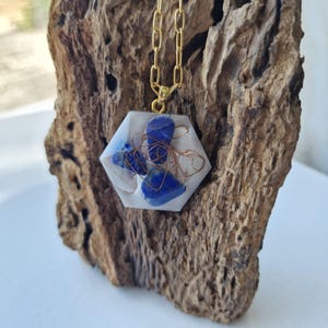 Pode incluir: Um colar com pingente hexagonal e corrente dourada. O pingente é feito de resina transparente com pedras azuis e detalhes em fio de cobre. O colar é exibido em um pedaço de madeira texturizada.