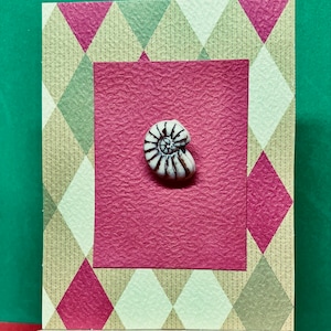May include: A white ceramic seashell with brown details is displayed on a red and white patterned card.