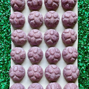 May include: A collection of twenty-four mauve-colored, spherical buttons with a detailed, textured floral design. The buttons are arranged in a grid on a light-colored cardboard backing, set against a backdrop of green tinsel.