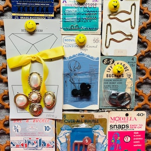 May include: A collection of vintage sewing notions, including zippers, hooks, snaps, buckles, and button covers. The items are displayed on a textured background and held in place with yellow smiley face magnets.