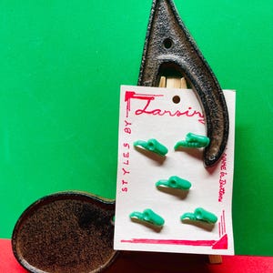 May include: A set of six green plastic buttons in a unique shape, displayed on a vintage card with the text "Lansing" and "Styles By". The card is held by a dark metal object, set against a red and green background.