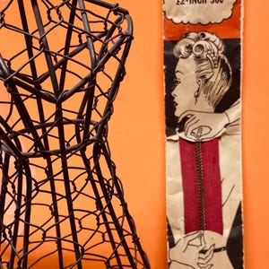 May include: A black wire dress form stands next to a vintage Ideal slide fastener package. The package features a red zipper illustration and the text "12-INCH 30c". The background is orange.
