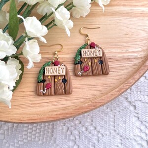 May include: A pair of gold earrings with a wooden door design. The doors are brown with a green vine and red flowers. Each door has a sign that says "Honey".