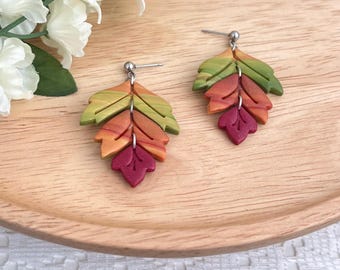Könnte beinhalten: Ein Paar ohrförmige Ohrringe in Herbstfarben. Die Ohrringe bestehen aus geschichtetem Polymer Clay, mit Grüntönen, Orange und Burgunderrot. Die Ohrringe liegen auf einem Holztablett mit weißen Blumen im Hintergrund.