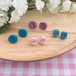 May include: A set of six colorful knotted earrings. The earrings are in three colors: teal, pink, and mauve. The earrings are displayed on a wooden surface.