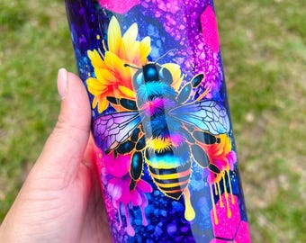 May include: A stainless steel tumbler with a colorful design featuring a bee, flowers, and honeycomb. The tumbler has a pink and purple glitter finish and is decorated with gold bee charms.