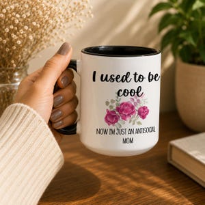 May include: A white ceramic mug with a black interior and handle, featuring the black text "I used to be cool" and "NOW I'M JUST AN ANTISOCIAL MOM" with a graphic of three pink roses and green leaves printed on the front surface of the mug.