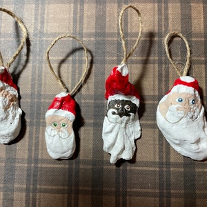 Könnte beinhalten: Vier handbemalte Austernmuschel-Ornamente mit Weihnachtsmann-Gesichtern. Jedes Ornament hat einen roten Hut und einen weißen Bart. Die Ornamente sind mit verschiedenen Gesichtszügen verziert, darunter blaue Augen, grüne Augen und ein schwarzes Gesicht.