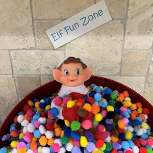 May include: A red bowl filled with colorful pom-poms and a smiling elf figurine. A sign above the bowl reads "Elf Fun Zone" in blue text. The pom-poms are various colors, including red, orange, yellow, green, blue, and purple.