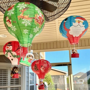 May include: A collection of colorful Christmas-themed hot air balloon lanterns. The lanterns feature festive designs like Santa Claus, reindeer, snowmen, and Christmas trees. Each balloon has a small basket with a plush toy inside.