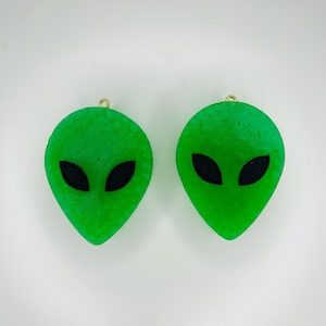 May include: Two green alien head-shaped ornaments with black eyes. The ornaments have a gold loop at the top for hanging.