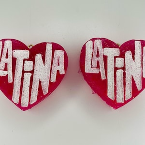 May include: Two pink heart-shaped resin earrings with the word "LATINA" written in white on each earring.