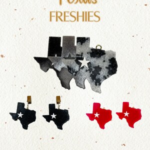 May include: Texas Freshies, a set of three Texas-shaped air fresheners. One is black with a white star, one is red with a white star, and one is a gray and white marble design with a white star.
