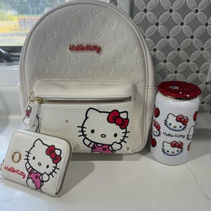 May include: White Hello Kitty backpack, wallet, and tumbler set. The backpack features embossed bow patterns and a front pocket. The wallet and tumbler have Hello Kitty graphics. The set includes a gold zipper and red accents.