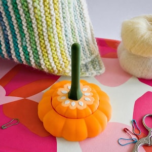 May include: A bright orange pumpkin-shaped yarn bowl with a dark green stem handle. The bowl features a white seed pattern around the centre stem. It is accompanied by a multi-coloured knitted fabric, a ball of cream yarn, and metal scissors.