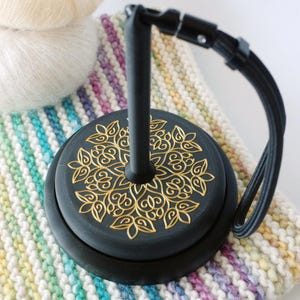 May include: A black yarn bowl stand featuring a circular base with an intricate gold floral mandala pattern and a central vertical post. A black elastic cord with a metal clasp is attached to the top of the post, extending downward to the side.