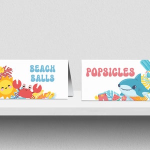 May include: Two white cards with colourful illustrations and text. The left card has a sun, crab, and palm leaves with the text "BEACH BALLS". The right card has a shark, watermelon, and popsicle with the text "POPSICLES".