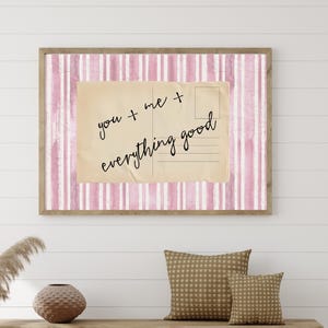 May include: A framed print with a pink and white striped background and a vintage postcard design. The postcard reads "you + me + everything good" in black script. The frame is a light brown color.